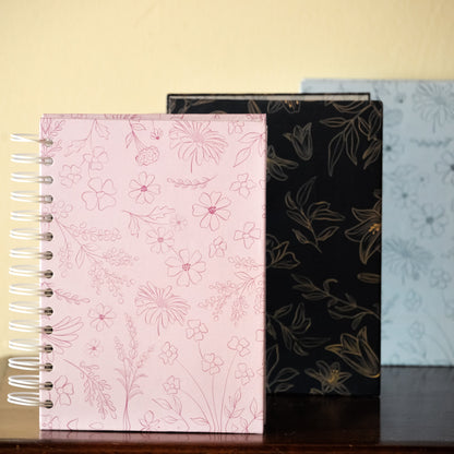 Three notebooks with floral patterns on a wooden surface against a beige wall.