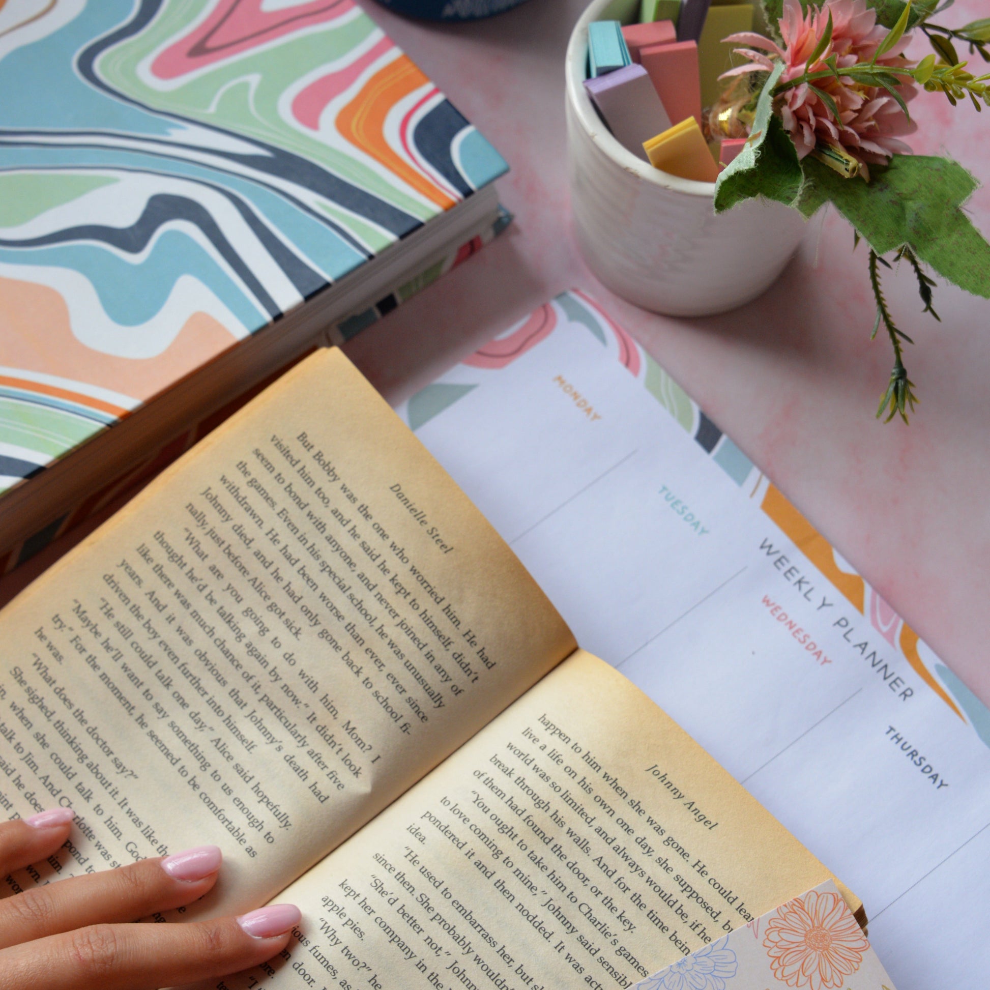 Open book with a hand on a pink surface, next to a colorful notebook, water bottle, and small plant.