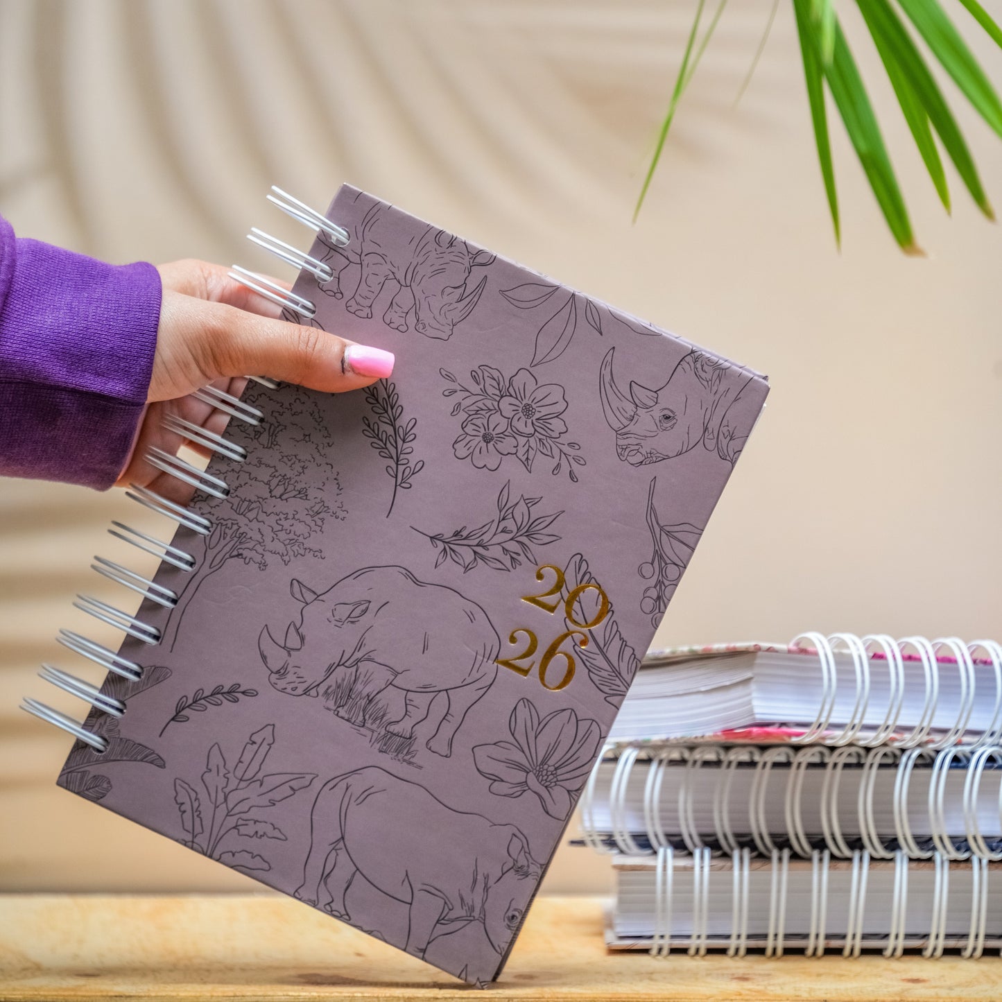 Hand holding a notebook with animal patterns and the number 2026 on a wooden surface with a plant in the background.