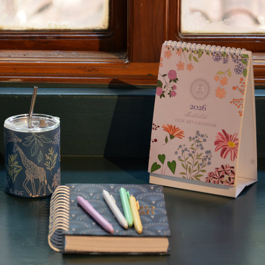 Notebook with colorful pens, candle, and floral-patterned calendar on a surface.