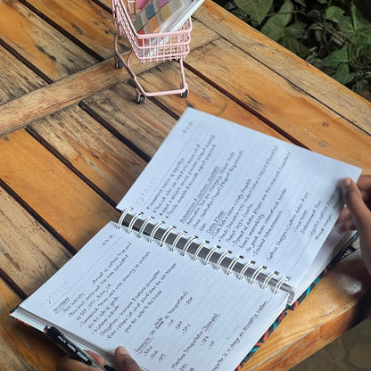Person holding an open notebook on a wooden table with a small shopping cart nearby.